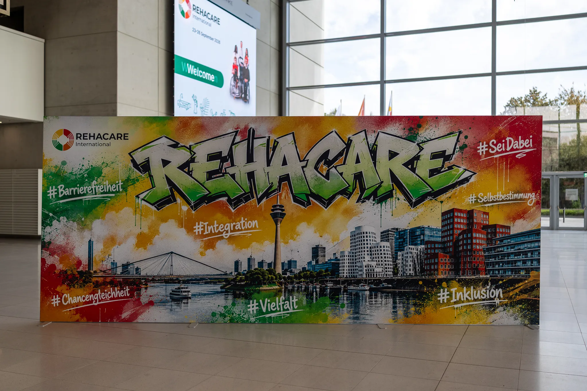 Bunte Fotowand mit REHACARE im Graffiti-Stil, der Düsseldorfer Skyline und Begriffen wie Barrierefreiheit, Selbstbestimmung und Inklusion.