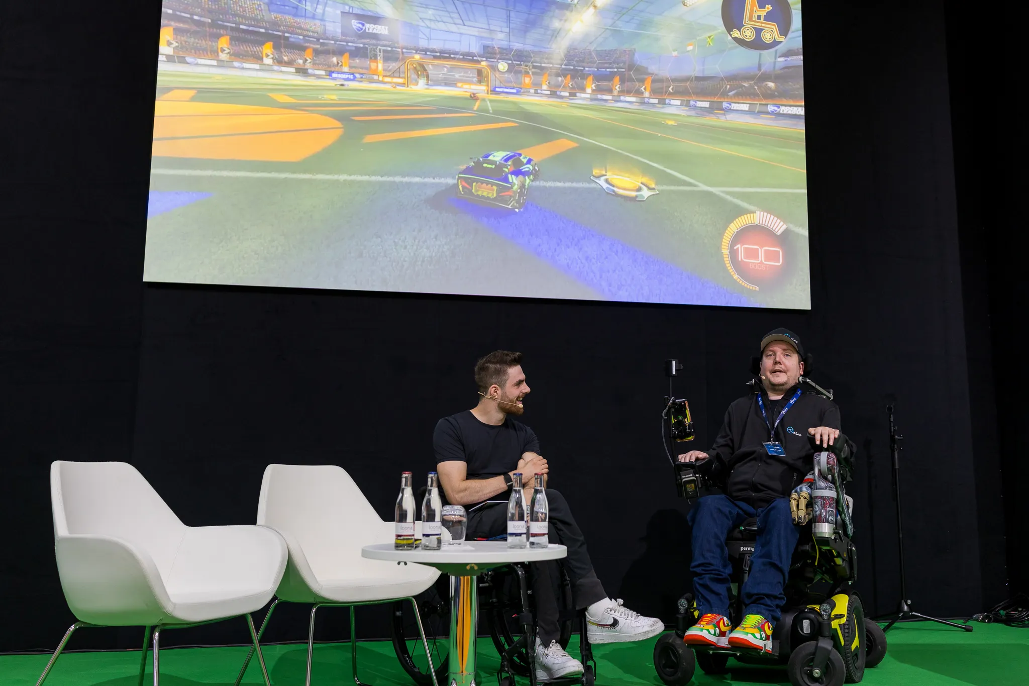Dennis Winkens sitzt rechts im Rollstuhl neben seinem Gesprächspartner; über ihnen läuft Rocket League auf einer Leinwand.