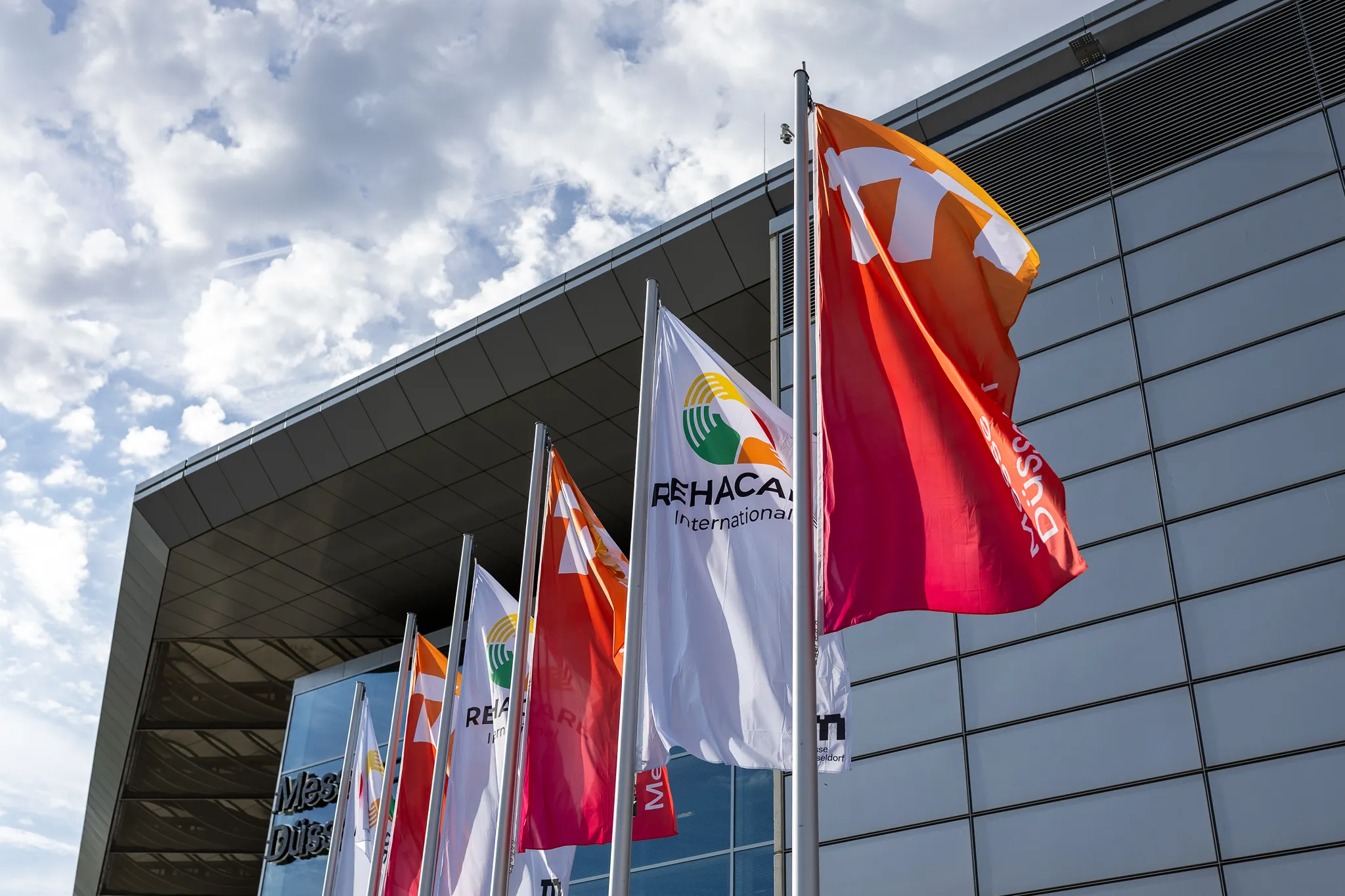 Weiße REHACARE-Flaggen und orange-rote Flaggen der Messe Düsseldorf vor der Fassade des Eingangsgebäudes.